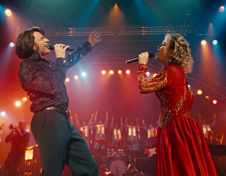 A man and woman perform on stage, their voices blending beautifully in a moving rendition of "Song Sung Blue." Cinematic lighting by Amy Vincent BSC enhances the captivating atmosphere.