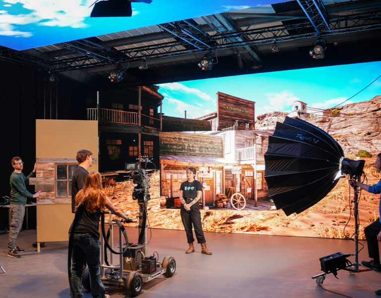 A film crew works on a Sony VP studio set with a large digital backdrop displaying a Western town scene. Several people, including Norwich University of the Arts students, handle equipment such as cameras, lighting, and set pieces.